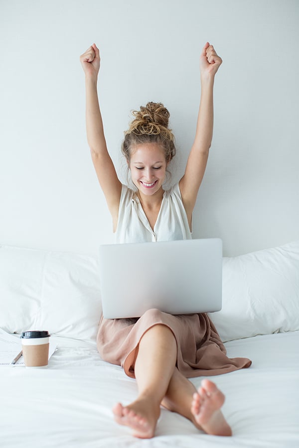 Woman siting on a bed with a laptop on her lap, and holding hands in the air - flexible working. 